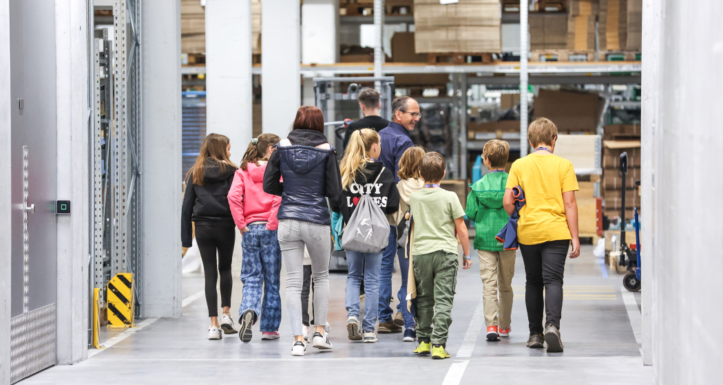 Bündner Mint-Woche 2025 Kindergruppe mit Begleitpersonen bei einer Betriebsbesichtigung während der Bündner MINT-Woche, unterwegs durch ein modernes Lager mit Regalen und Materialien.