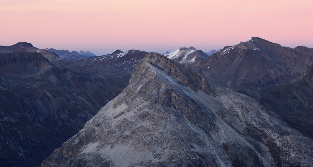 Blick auf den Piz Alv in den Bündner Alpen bei Abendstimmung, mit felsigem Gipfel, karger Berglandschaft und rosa gefärbtem Himmel im Hintergrund.