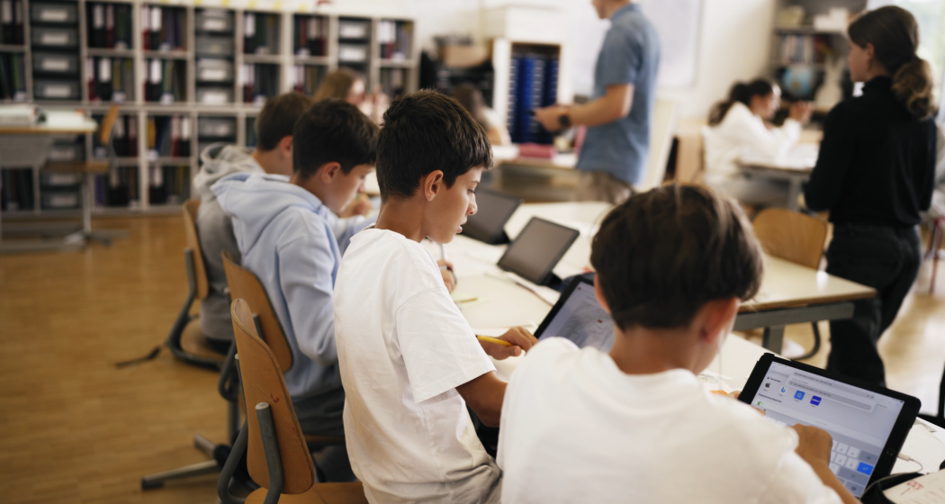 Schülerinnen und Schüler arbeiten im Klassenzimmer mit Tablets, während eine Lehrperson im Hintergrund unterrichtet