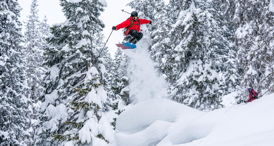 Skifahrer in roter Jacke springt über eine Schneewechte in verschneiter Winterlandschaft mit schneebedeckten Tannen.