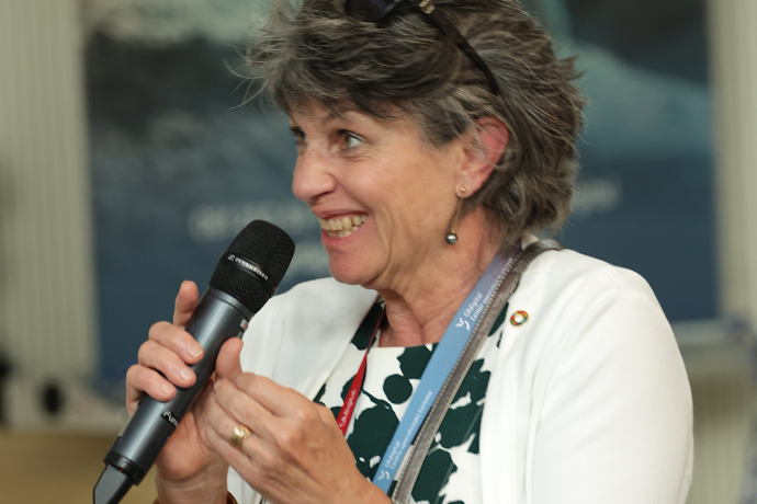 Barbara Haller Rupf spricht lächelnd mit einem Handmikrofon an der Generalversammlung von graubünden digital 2026. Sie trägt eine weisse Jacke und mehrere Lanyards, im Hintergrund ist ein grossformatiges Kunstwerk zu sehen.