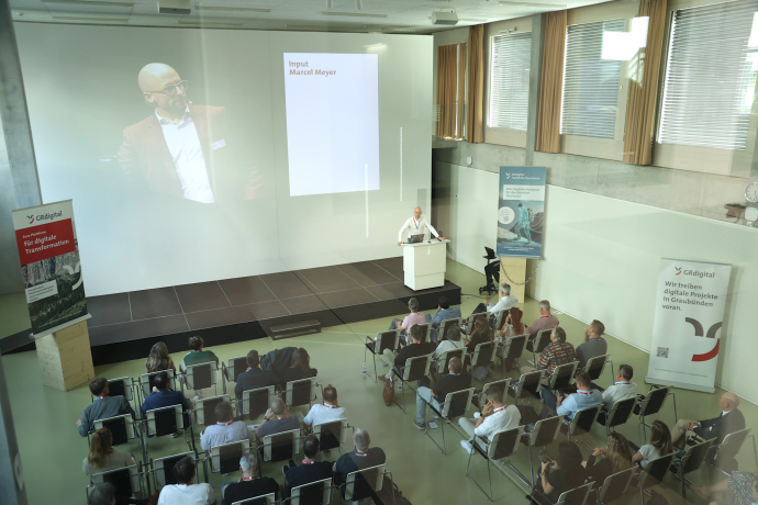 Marcel Meyer hält seinen Inputvortrag an der Generalversammlung von graubünden digital 2026. Auf dem Präsentationsscreen im Hintergrund ist sein Name und das Wort «Input» zu lesen. Links und rechts stehen Roll-up-Banner von GRdigital, das Publikum sitzt dem Referenten gegenüber.