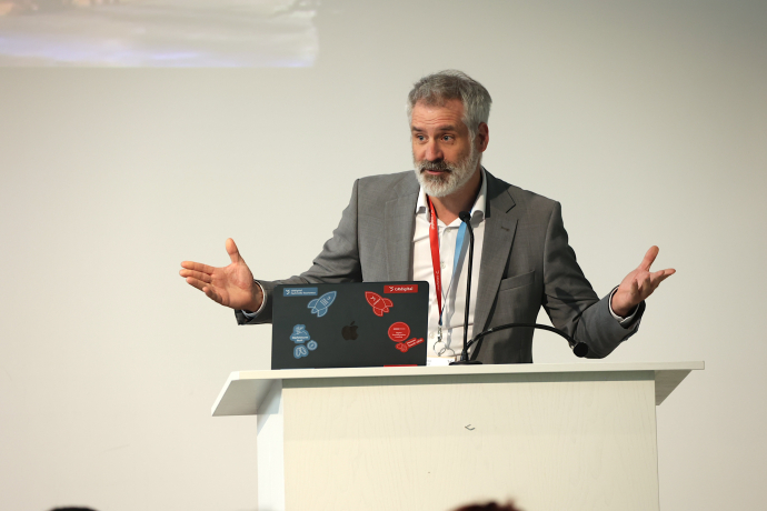 Nikolaus Schmid referiert lebhaft mit ausgebreiteten Armen am Rednerpult der Generalversammlung von graubünden digital 2026. Vor ihm steht ein Laptop mit GRdigital-Stickern, er trägt ein rotes Lanyard.