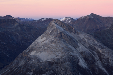 Blick auf den Piz Alv in den Bündner Alpen bei Abendstimmung, mit felsigem Gipfel, karger Berglandschaft und rosa gefärbtem Himmel im Hintergrund.