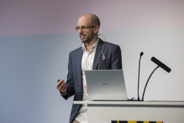 Marcel Meyer steht auf einer Bühne hinter einem Rednerpult und spricht mit Headset-Mikrofon vor Publikum. Vor ihm steht ein offener Laptop.