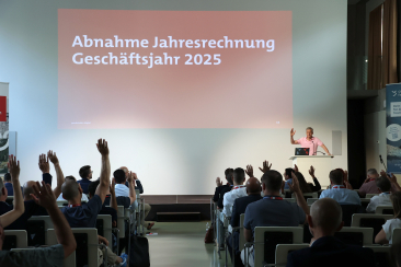 Abstimmung an der Generalversammlung von graubünden digital 2026: Die Teilnehmenden heben die Hand zur Annahme der Jahresrechnung 2025, während dJon Erni am Rednerpult auf der Bühne ebenfalls die Hand hebt. Im Hintergrund ist die Präsentationsfolie mit der Aufschrift «Abnahme Jahresrechnung Geschäftsjahr 2025» zu sehen.
