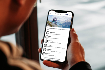Person hält Smartphone in der Hand und nutzt die Engadin-App mit Buchungsoptionen für Bergbahn-Tickets, Bike-Miete und Aktivitäten, im Hintergrund Bergpanorama.