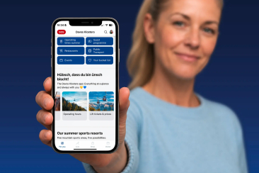 Frau hält Smartphone mit geöffneter Davos Klosters App, die Menüpunkte wie Öffnungszeiten, Gästeprogramm, Restaurants, ÖV, Events und Bucket List anzeigt.