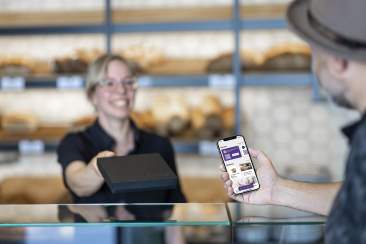 Bäckerei-Verkäuferin reicht einem Kunden über die Theke eine schwarze Schachtel. Der Kunde hält ein Smartphone mit geöffneter App (Bereiche «Vereine» und «Aktionen»).